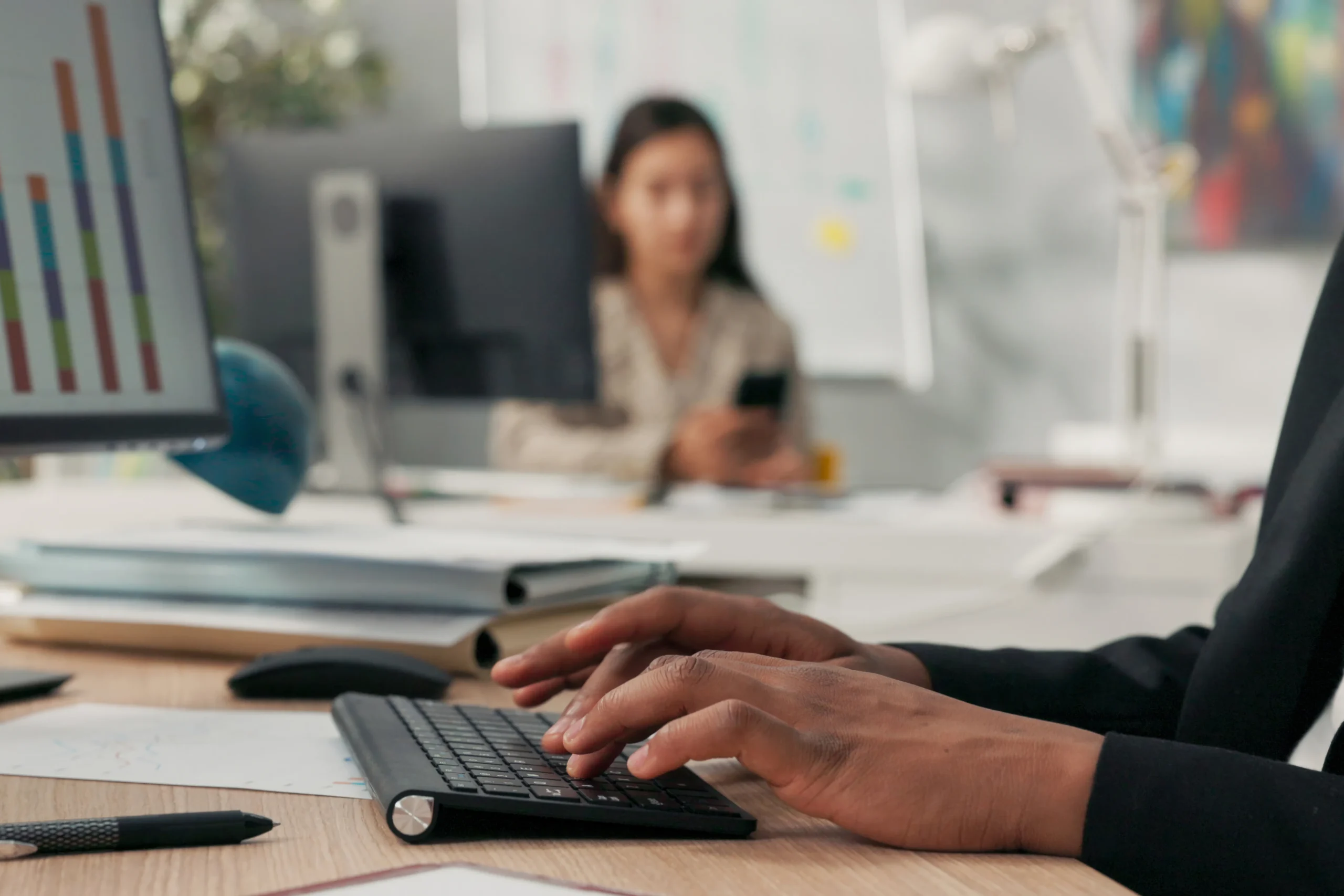 A man works at a desk on a computer in the foreground, and a woman works at a desk in the background