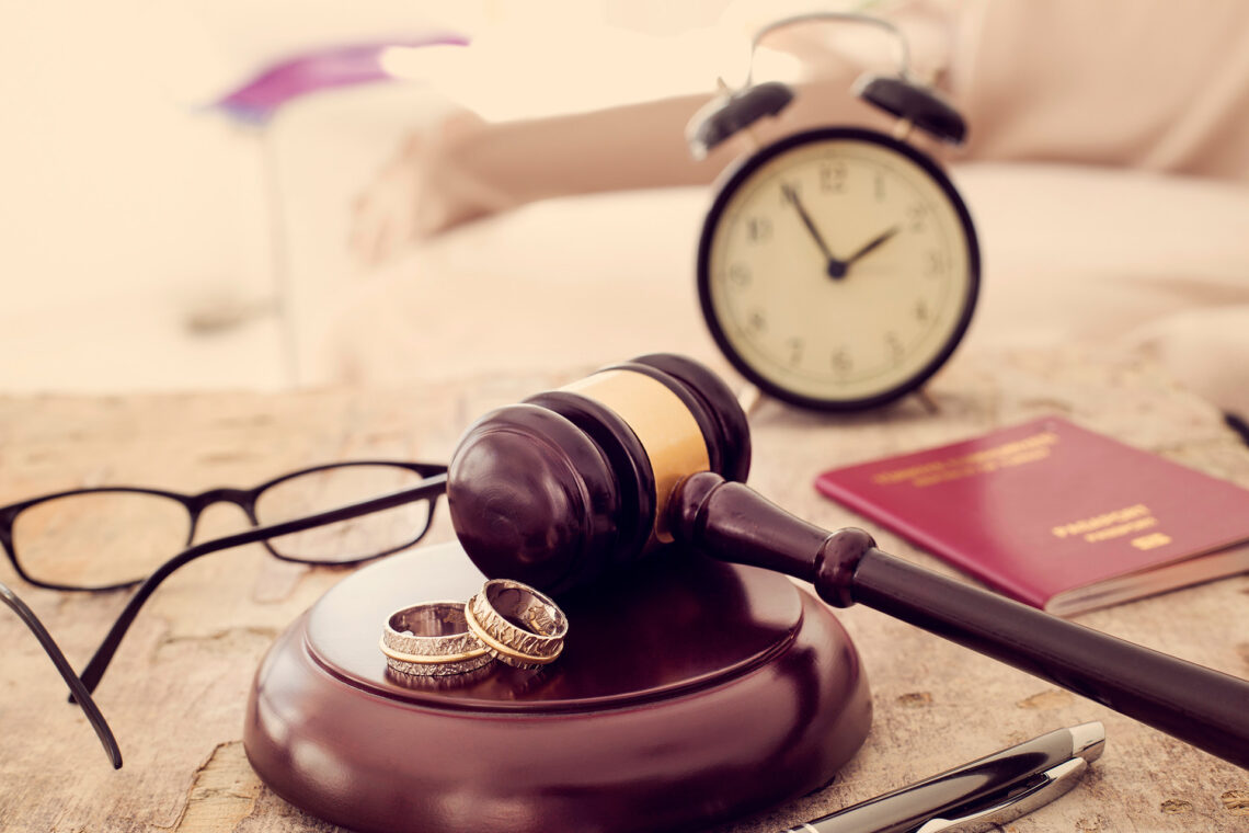 A gavel on a desk with two wedding rings, glasses, a clock, a pen, and a passport. The scene conveys themes of legal decisions and divorce.