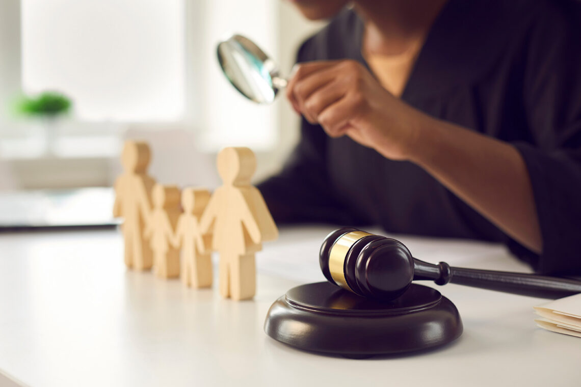 A wooden carved family ornament on a desks with a judge sitting at the desk with a magnifying glass in their hard looking at the ornament.