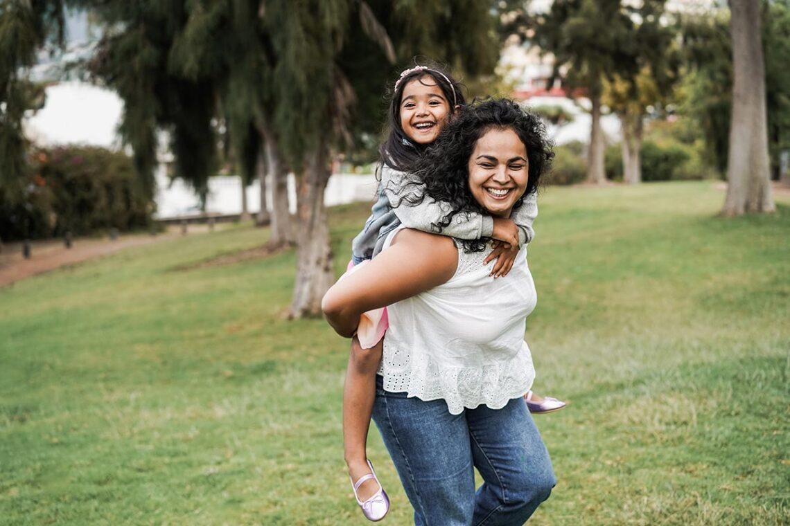 A smiling woman gives a young girl a piggyback ride through a grassy park.