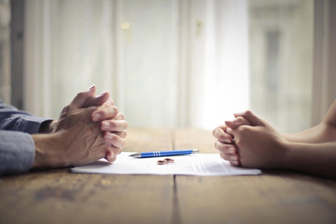 A table with a divorce agreement and wedding rings on top with the hands of two people on either side of the table.